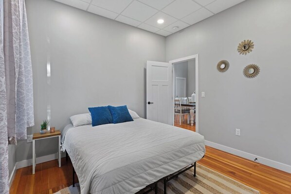Un dormitorio con cama, mesita de noche, una silla y una puerta que da a otra habitación.