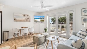 A modern living room with a ceiling fan, a dining area with white chairs and a table, and a view of the outdoors through large sliding doors. - Miramar Beach Condo: Pool, Hot Tub, Beach (Miramar Beach)