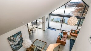 A spacious loft with a high ceiling, a staircase, a dining area, and a large window overlooking a body of water. - Kessock (Windermere)