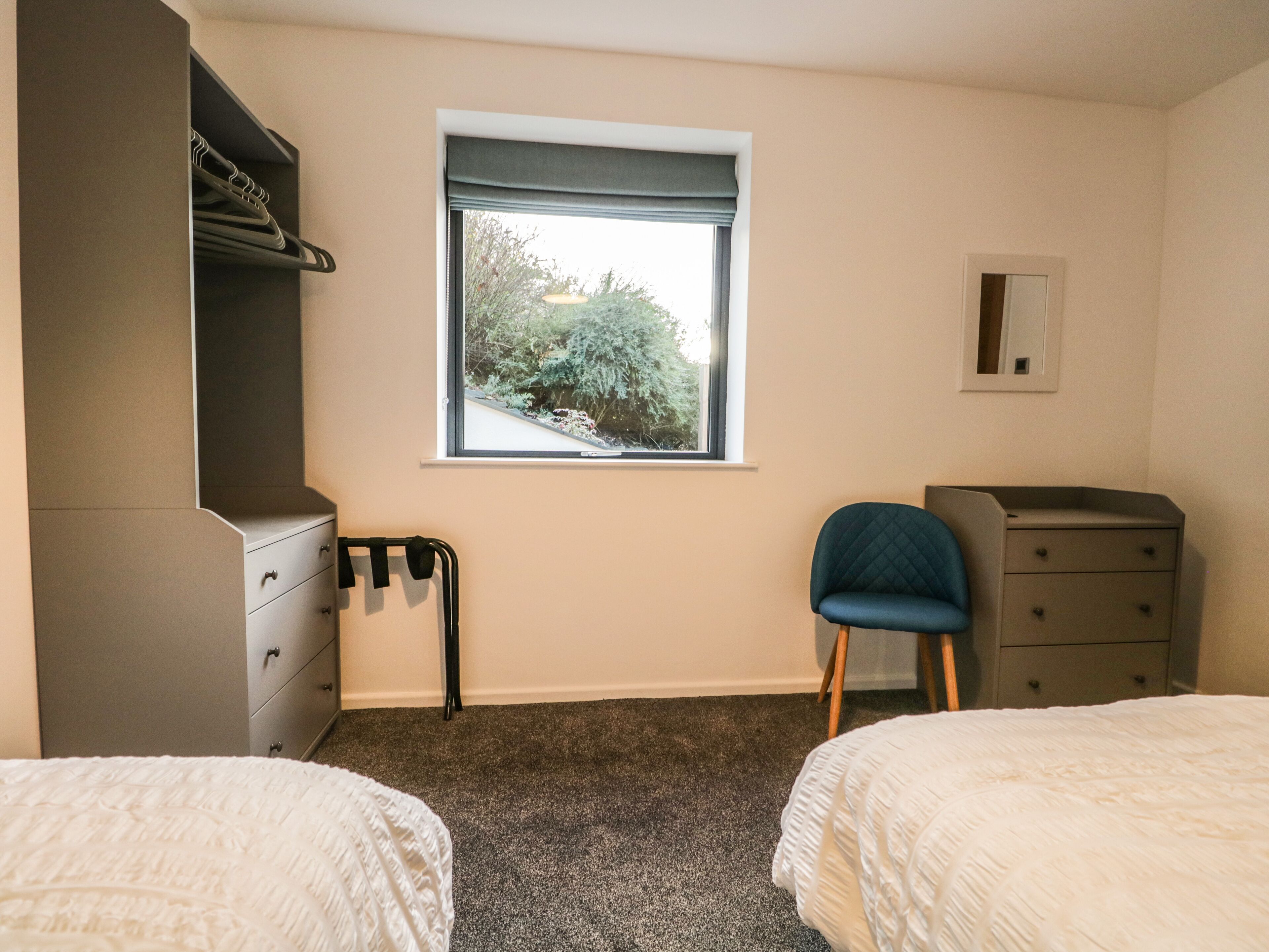 A bedroom with two beds, a window with a view of greenery, a wardrobe, and a chair.