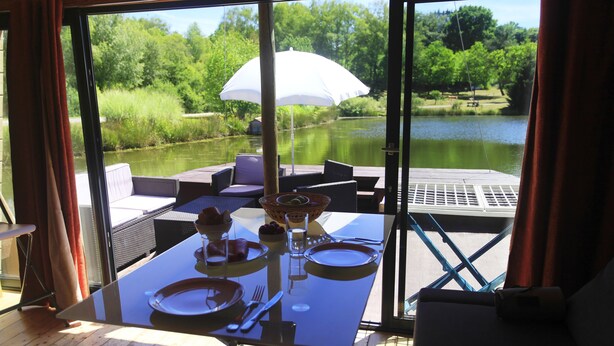 A dining table set with plates, cutlery, and a glass of water, overlooking a calm lake and lush greenery. - Toue cabanée (Chamberet)