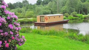A houseboat with a small deck and a cabin, floating on a calm river surrounded by greenery. - Toue cabanée (Chamberet)