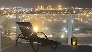 Ein Balkon mit Liegestuhl und Laterne mit Blick auf eine nächtliche Stadtlandschaft.