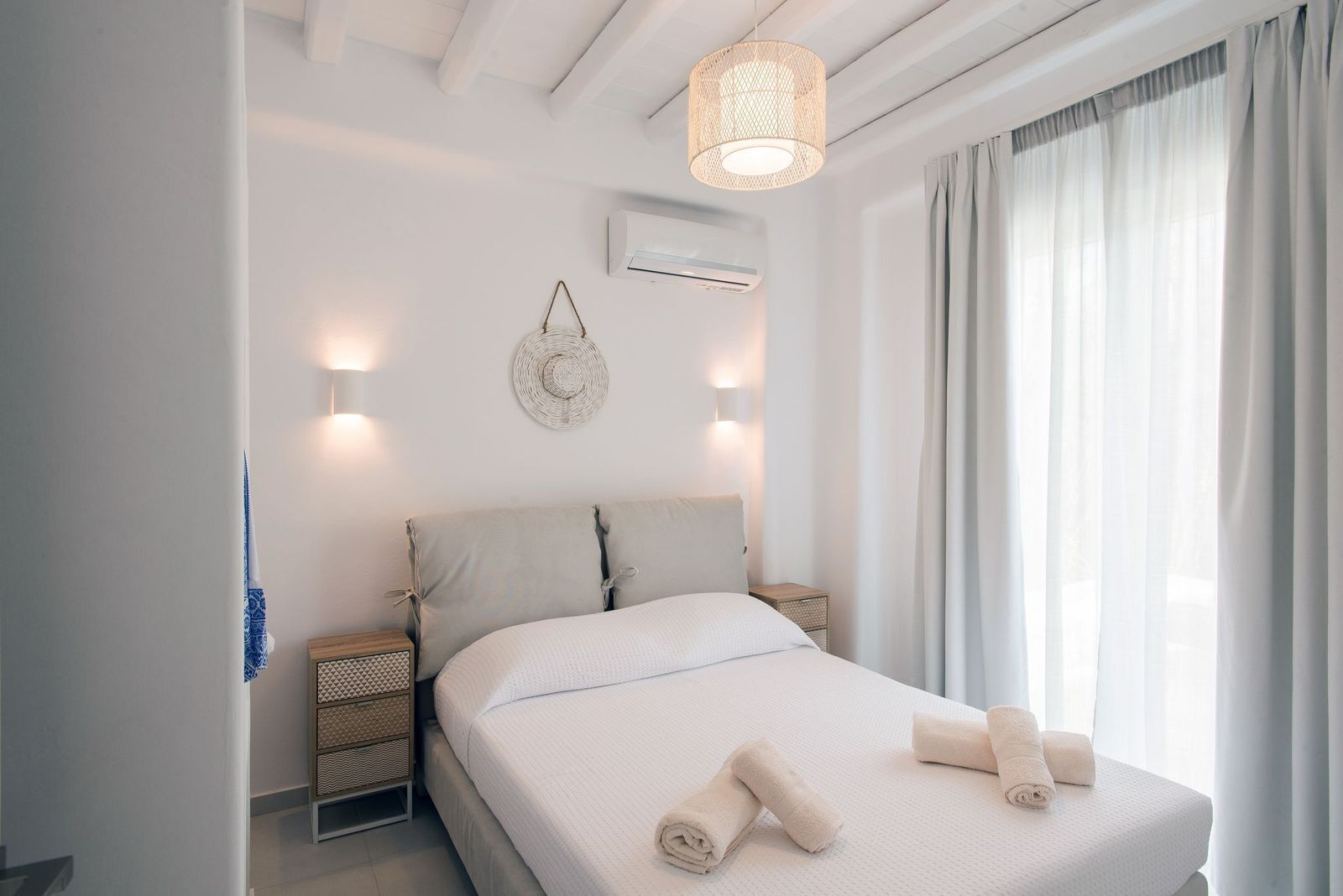 A modern bedroom with a large bed, two bedside tables, a hanging light fixture, and a window with curtains.