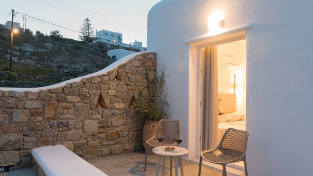 A stone wall with a bench, two wicker chairs, and a table with a plate and wine glasses. - Desire Mykonos Apartments (Mykonos)