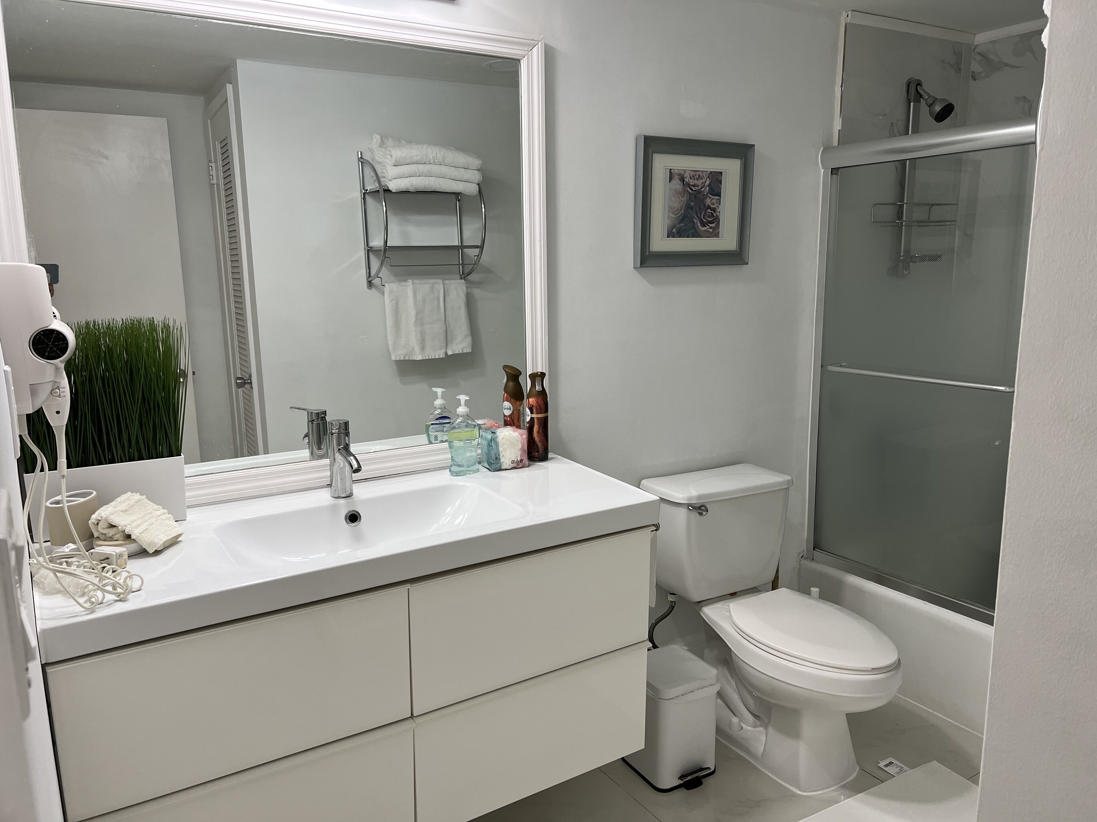 Ein modernes Badezimmer mit einem weißen Waschbecken, Spiegel und WC. Es gibt einen Duschbereich mit einer Glastür und ein gerahmtes Bild an der Wand.
