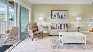 Un salon avec un fauteuil beige, un canapé beige, une table basse blanche et un tapis à motifs. Il y a un tableau au mur et deux lampes sur des tables d’appoint.