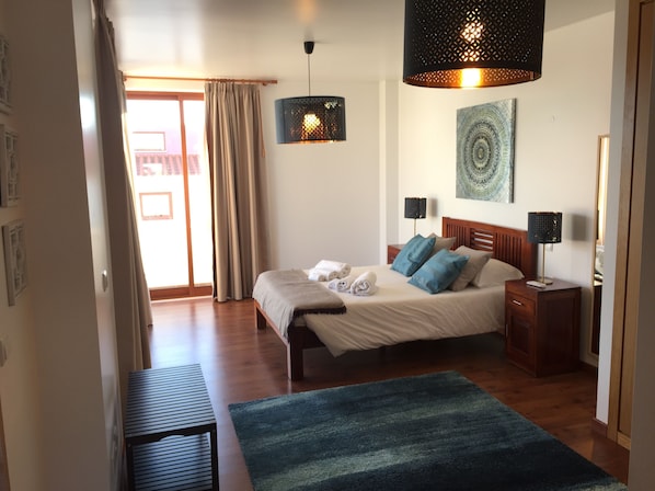 A hotel room with a bed, bedside tables, a blue rug, a hanging light fixture, and a painting on the wall.
