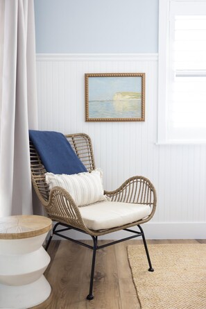 A wicker chair with a cushion and a blue throw blanket, positioned next to a white round table with a woven top.