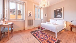 A bedroom with a bed, a desk, and a chair, a chandelier, and a window with a view of buildings. - GuestReady - Ray of Light (Lisbon)