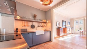 A modern kitchen with grey cabinets, a stainless steel countertop, and a wooden floor. There is a dining area with a table and chairs, and a large window providing natural light. - GuestReady - Ray of Light (Lisbon)