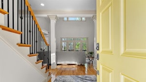 A staircase with wooden handrails and black balusters, a marble-patterned rug, a yellow door, and a window with a view of greenery.