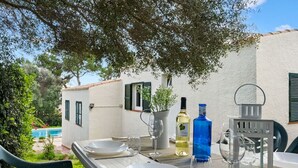 Un espace extérieur aménagé pour les repas, avec une table dressée pour deux personnes, une bouteille de vin blanc et une bouteille bleue, probablement d’huile d’olive, sur une terrasse donnant sur une piscine.