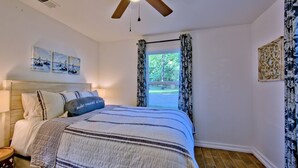 Ein Schlafzimmer mit Bett, Deckenventilator, Fenster mit Vorhängen und Bildern an den Wänden.