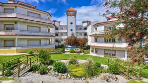 A courtyard with a central tower, surrounded by multi-story buildings with balconies and a small pond. - Praia Del Rey - Beach/Pool/Golf/Adventure (Amoreira)