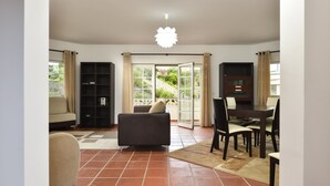 A spacious living room with a sofa, a dining table with chairs, a bookshelf, and a sliding glass door leading to a balcony. - Praia Del Rey - Beach/Pool/Golf/Adventure (Amoreira)