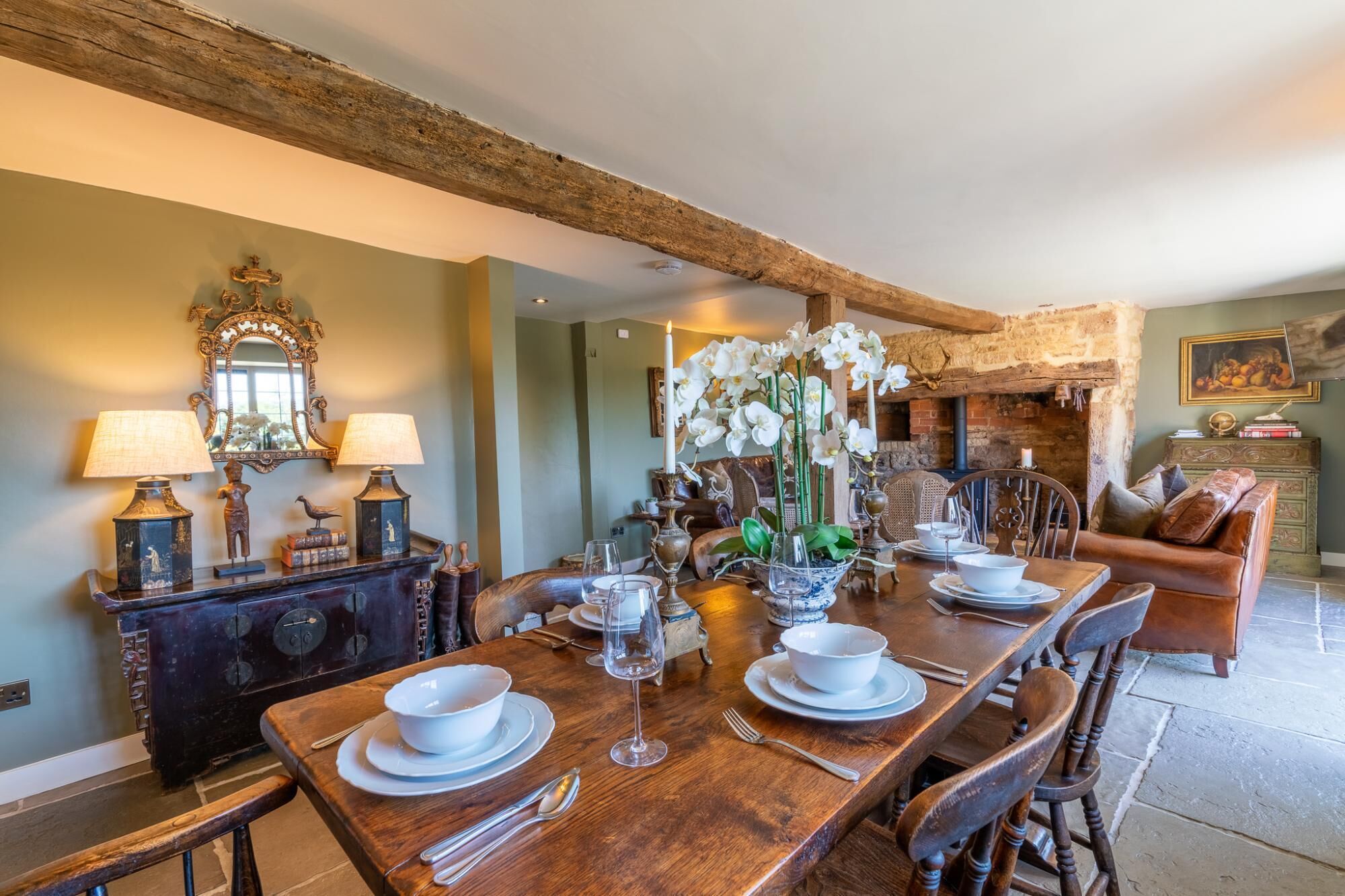 A dining area with a wooden table set for four, surrounded by chairs. There is a mirror on the wall, a fireplace, and a sofa in the background.