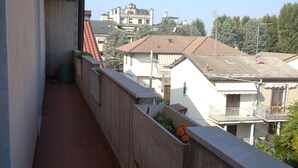 Uma varanda estreita com chão de azulejos, grades de concreto e vista para edifícios e árvores vizinhos.