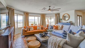 A spacious living room with a large orange sofa, a blue sofa, and a ceiling fan. There are large windows with curtains, a wooden cabinet, and a round mirror on the wall. - Best Luxury Riverfront Home w/ Private Pool & Dock (Bullhead City)
