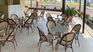 Un espacio de estar al aire libre techado con sillas y mesas de mimbre, piso de baldosas y vista a la calle.