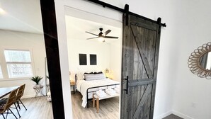 Una habitación con cama, ventilador de techo, puerta de madera y una zona de comedor con sillas y mesa.