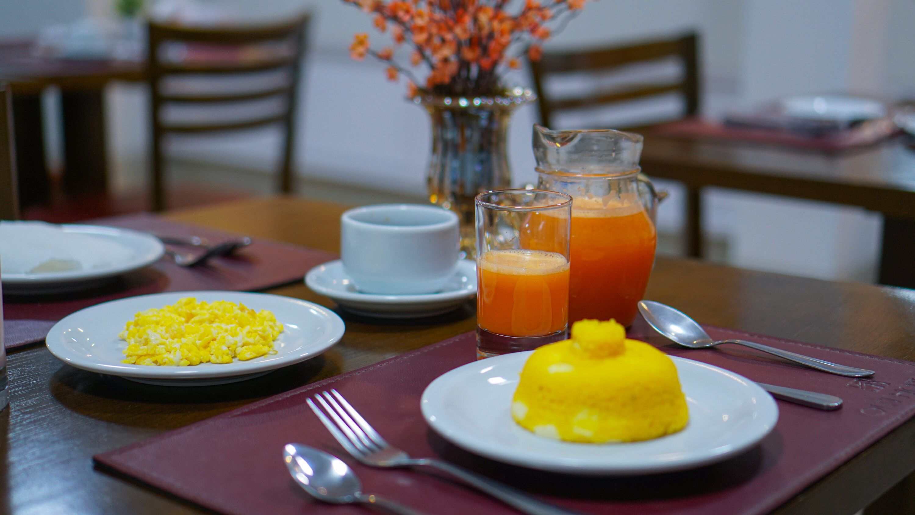 A breakfast setting with plates of scrambled eggs and a yellow dessert, a pitcher of orange juice, and a cup of coffee on a wooden table.