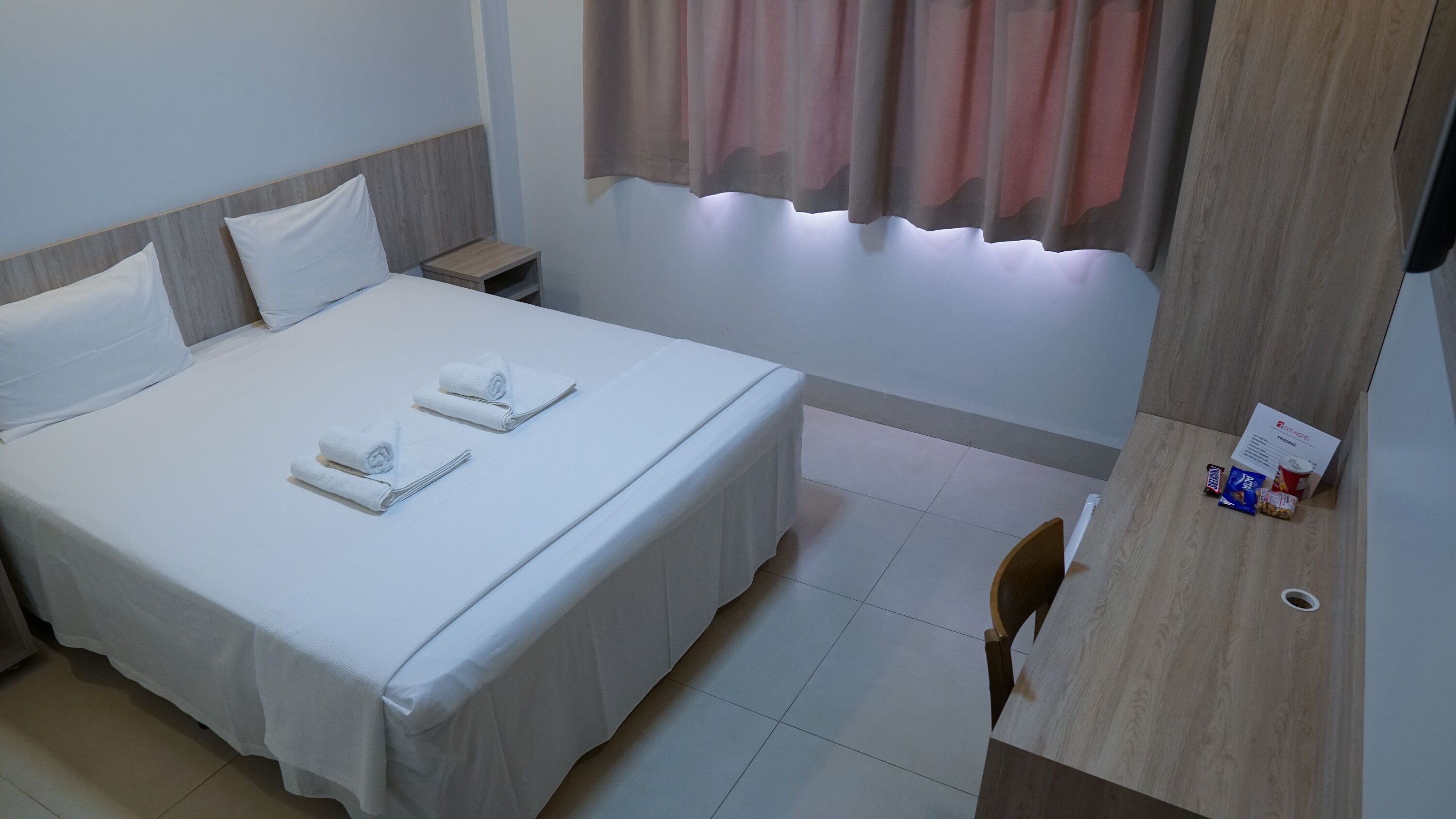 A hotel room with a bed, two pillows, a nightstand, a desk with a chair, a small table with snacks, and a window with curtains.