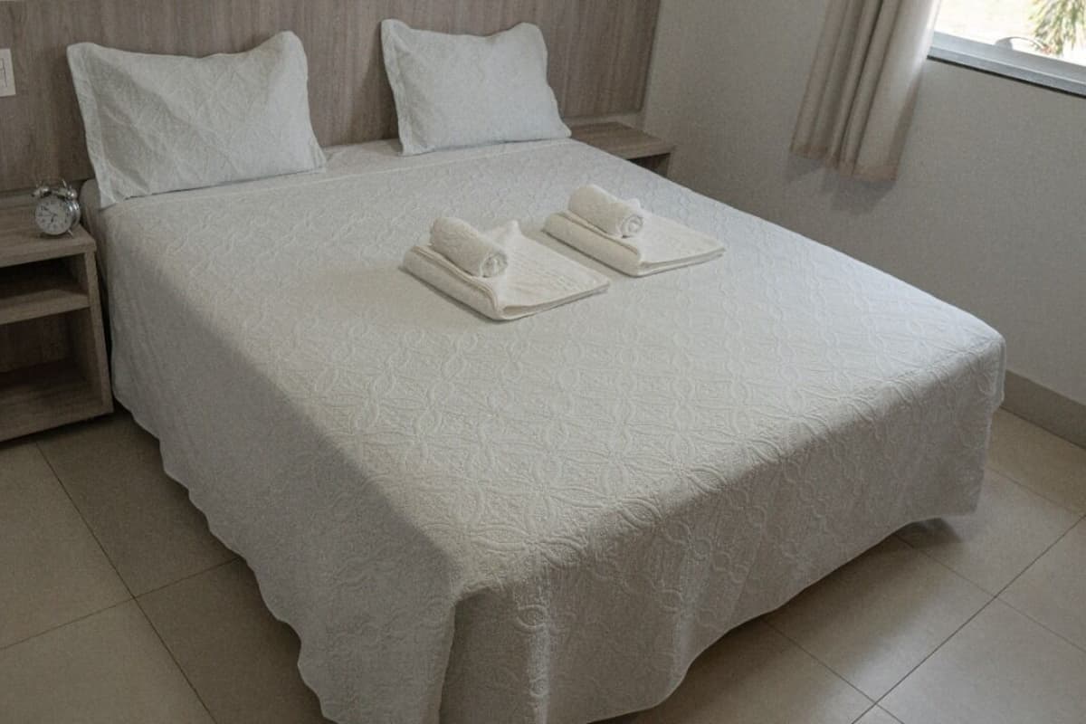 A neatly made bed with two pillows and two folded towels, a bedside table with a phone and an alarm clock, and a nightstand with a lamp.