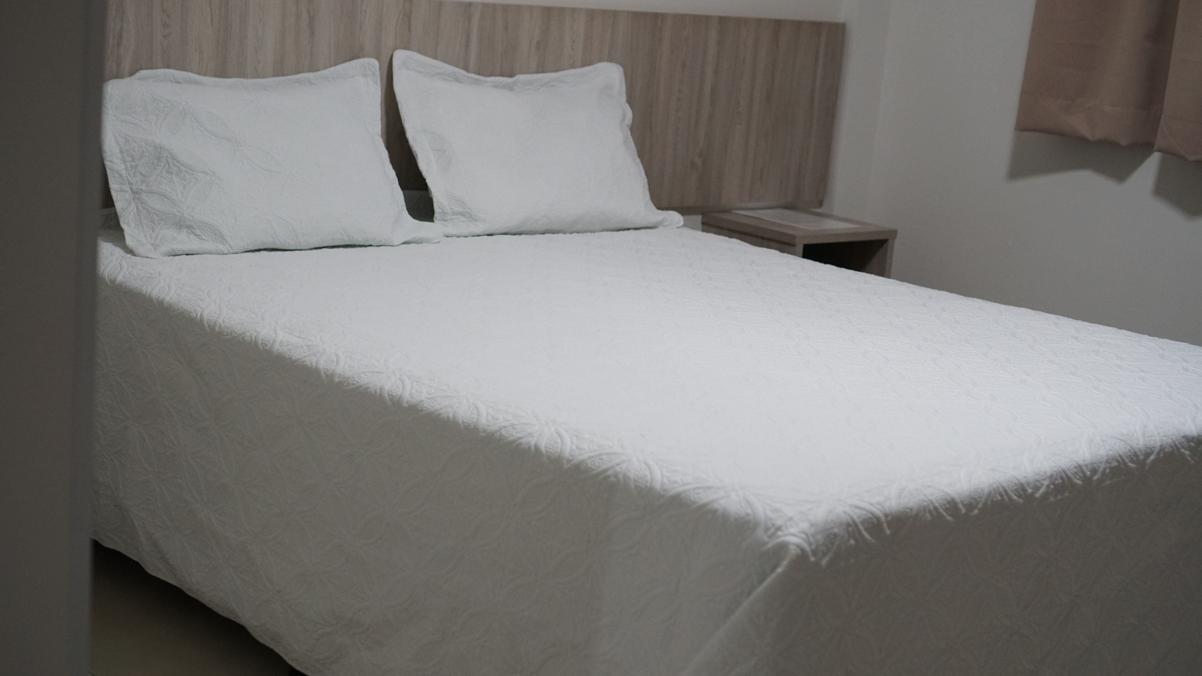 Una cama bien hecha con cubrecama y almohadas blancas, cabecera de madera y una mesita de noche con lámpara.