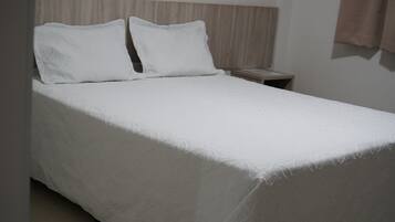 Una cama bien hecha con cubrecama y almohadas blancas, cabecera de madera y una mesita de noche con lámpara.