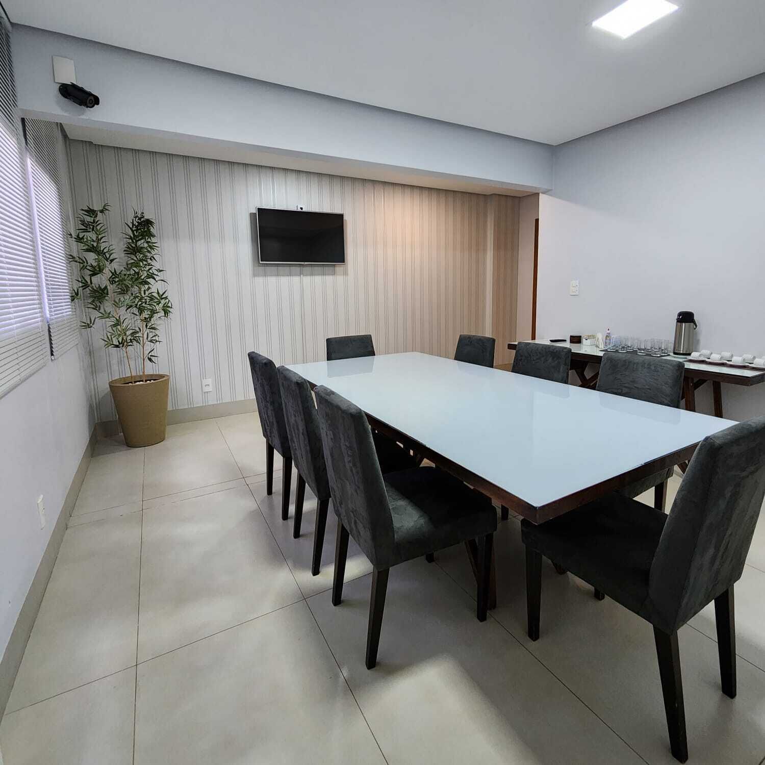 Una sala conferenze con un lungo tavolo, sedie, una pianta in vaso, una TV e un angolo caffè.