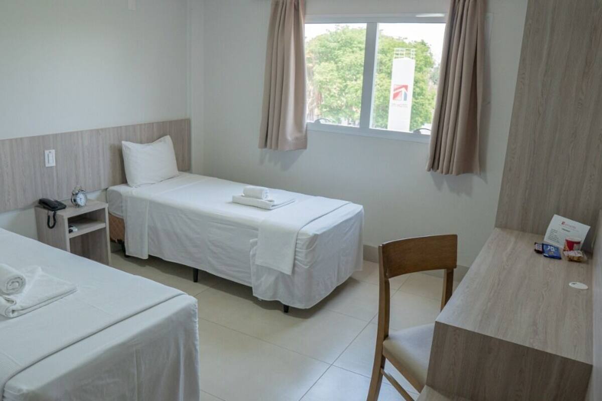 A hotel room with two single beds, a desk, a chair, a nightstand with a phone, and a window with curtains.