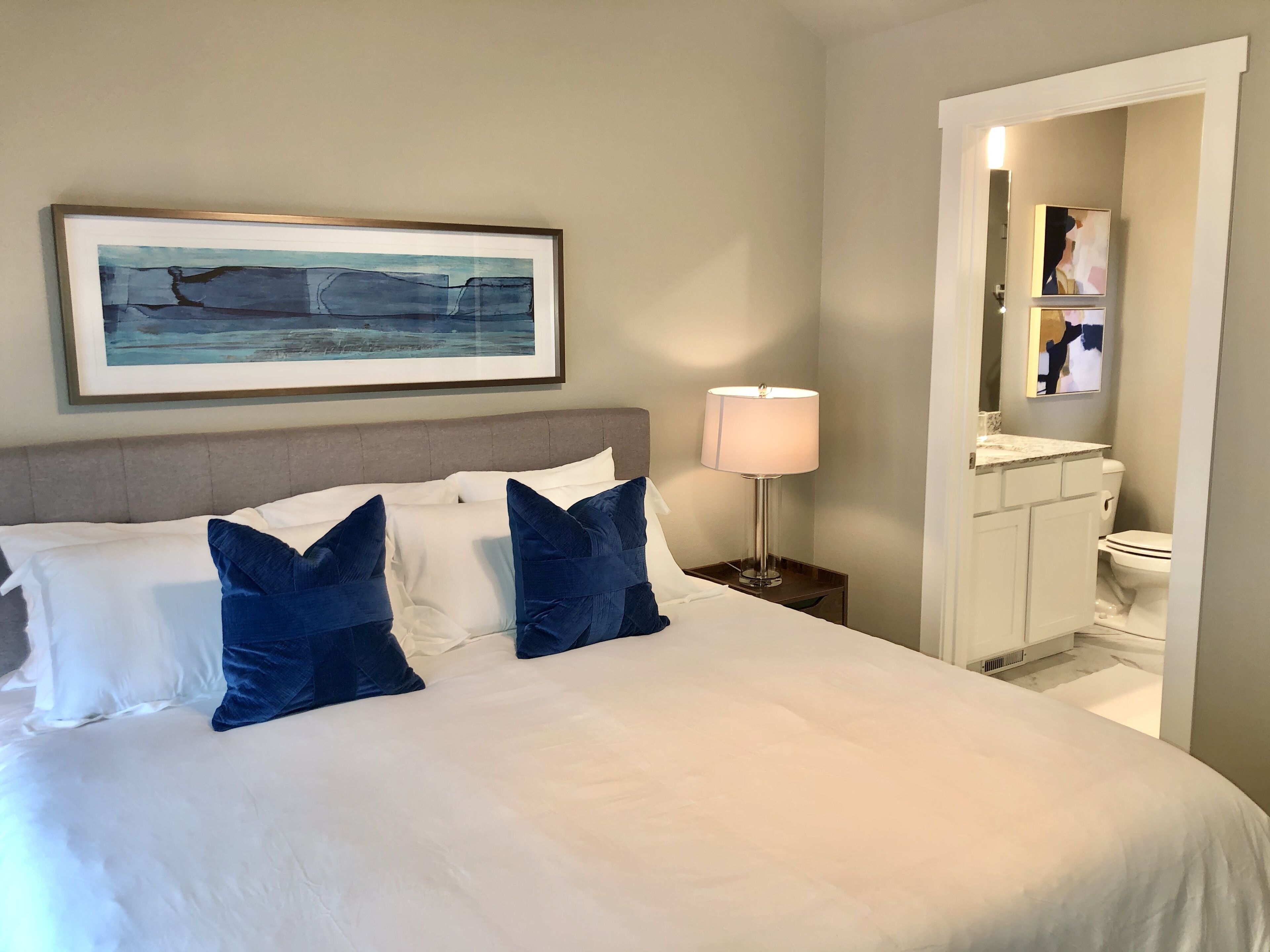 A hotel room with a bed, a nightstand, a lamp, a framed artwork, and a bathroom visible through an open door.