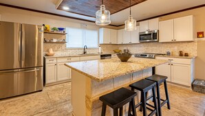 Une cuisine moderne dotée d’un grand îlot central, d’appareils électroménagers en acier inoxydable et de luminaires suspendus.