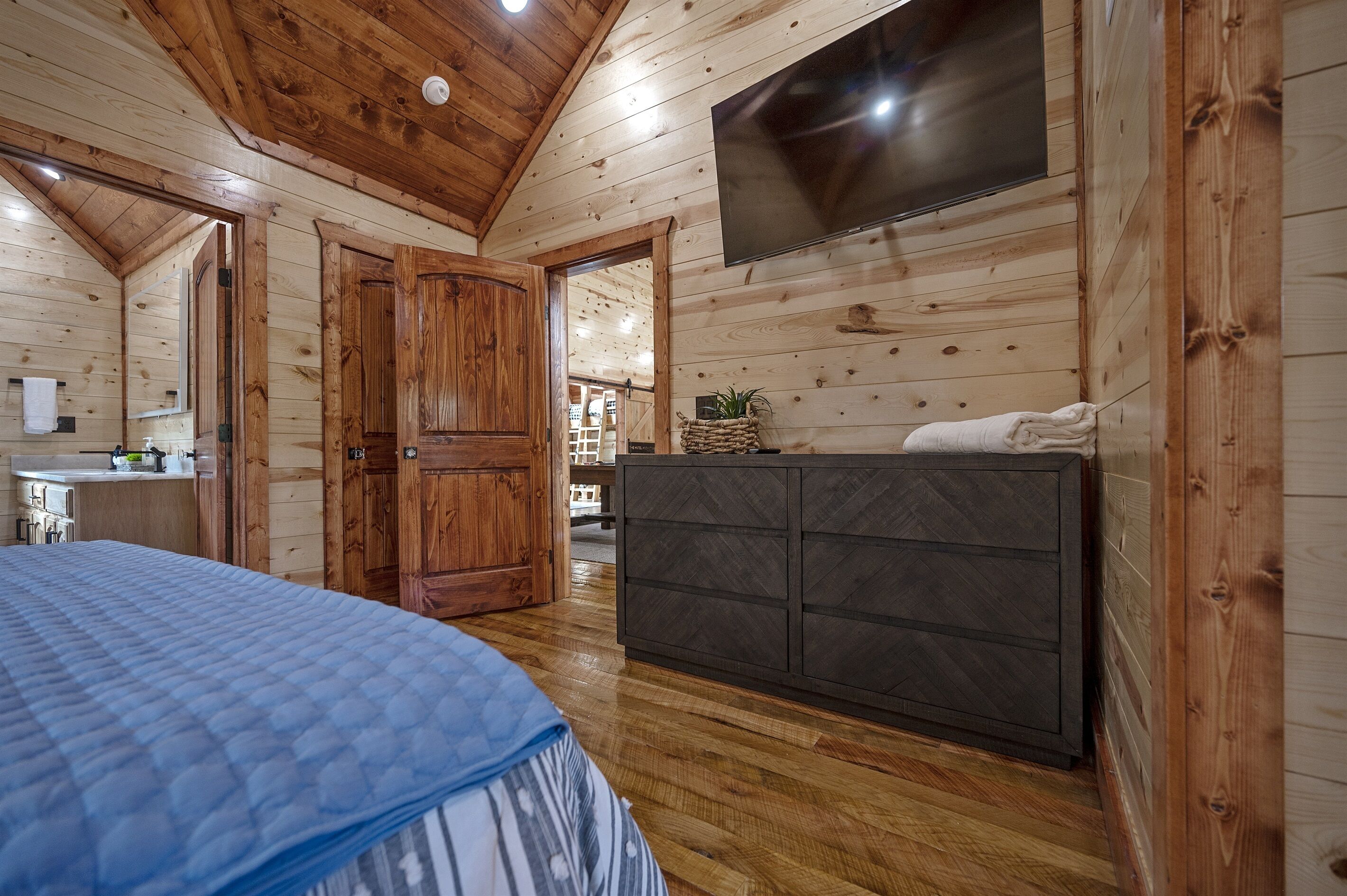 A wooden cabin interior with a bed, a dark wooden dresser, a TV mounted on the wall, and a kitchen area visible through an open door.