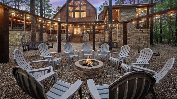 Una cabaña de troncos con chimenea de piedra rodeada de sillas Adirondack, situada en una zona boscosa.