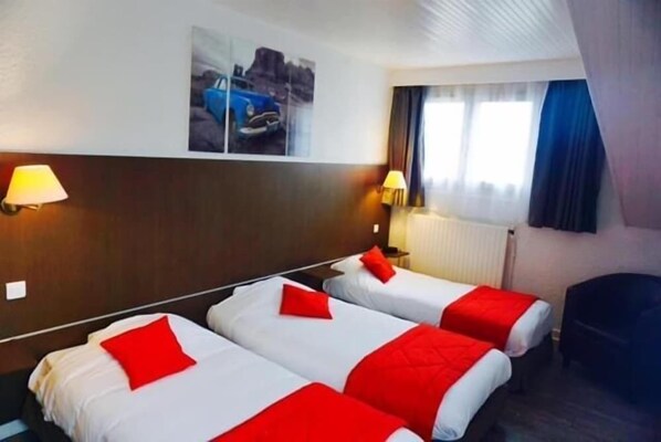 Habitación de hotel con dos camas, cada una con ropa de cama roja y blanca, y una mesita de noche con lámpara. Se observa una ventana con cortinas, una obra de arte en la pared y un radiador.
