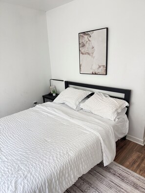 Een slaapkamer met een bed, een nachtkastje, een lijst met een foto aan de muur en een kamerkleed met een patroon.