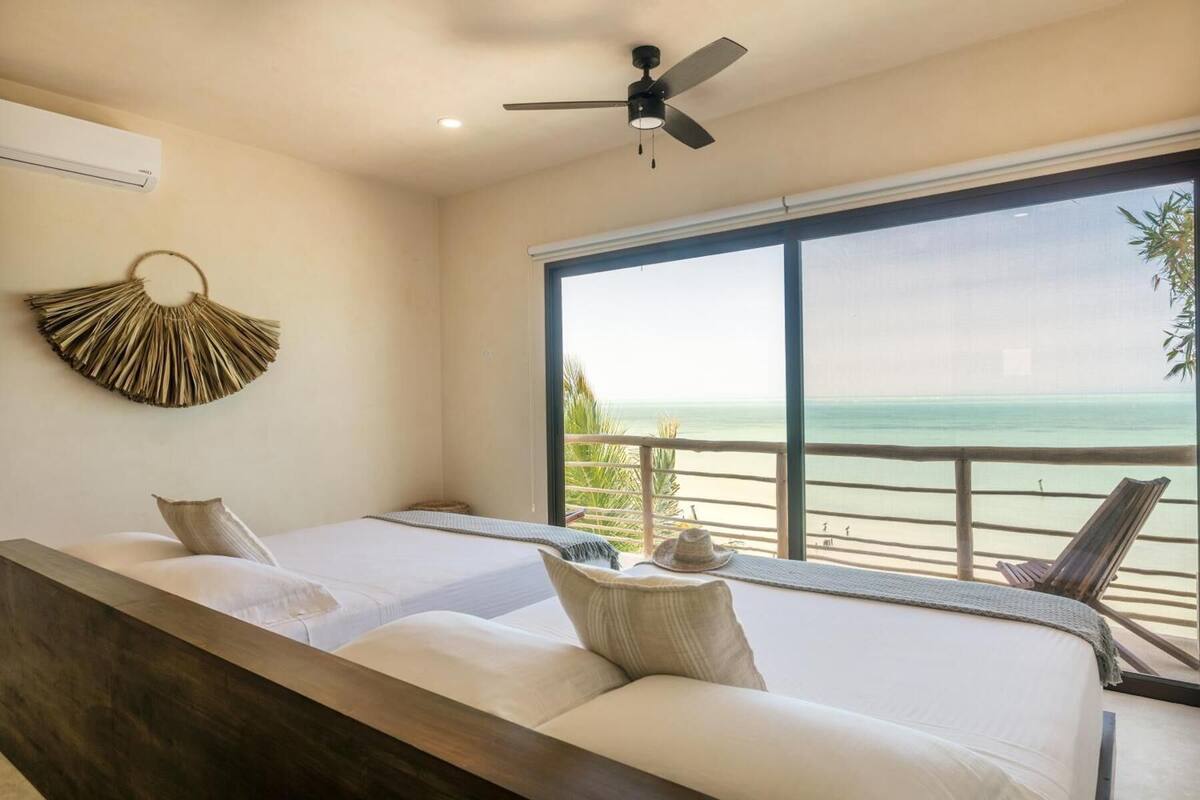 Ein Zimmer mit zwei Betten, einem Deckenventilator, einem großen Fenster mit Blick auf den Strand und einem Wanddekor.