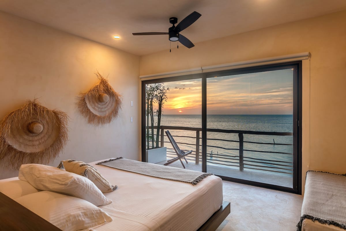 Un dormitorio con cama, almohadas, ventilador de techo y un ventanal con vistas al océano y al atardecer.