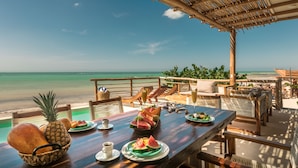 A wooden table with plates of food, a pineapple, and a basket of bread, overlooking a beach with a clear sky. - Hotel Boutique Casa Muuch Holbox (Isla Holbox)