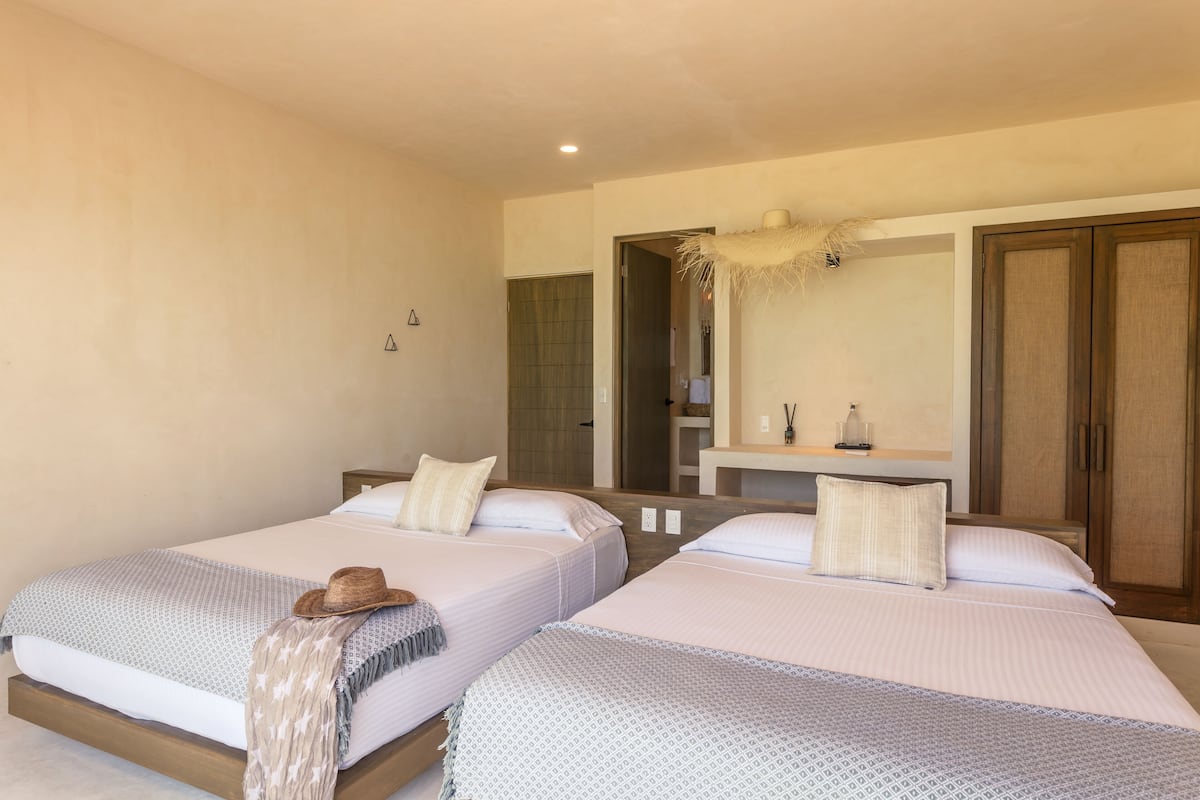 Habitación de hotel con dos camas, un espejo grande, armario de madera y un baño visible a través de una puerta abierta.