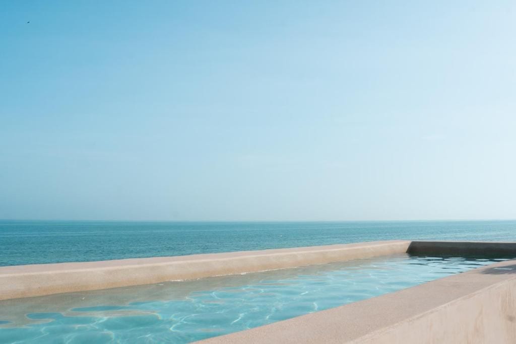 Vista despejada de una piscina con borde arenoso y superficie de agua tranquila, que da a una playa con horizonte claro.