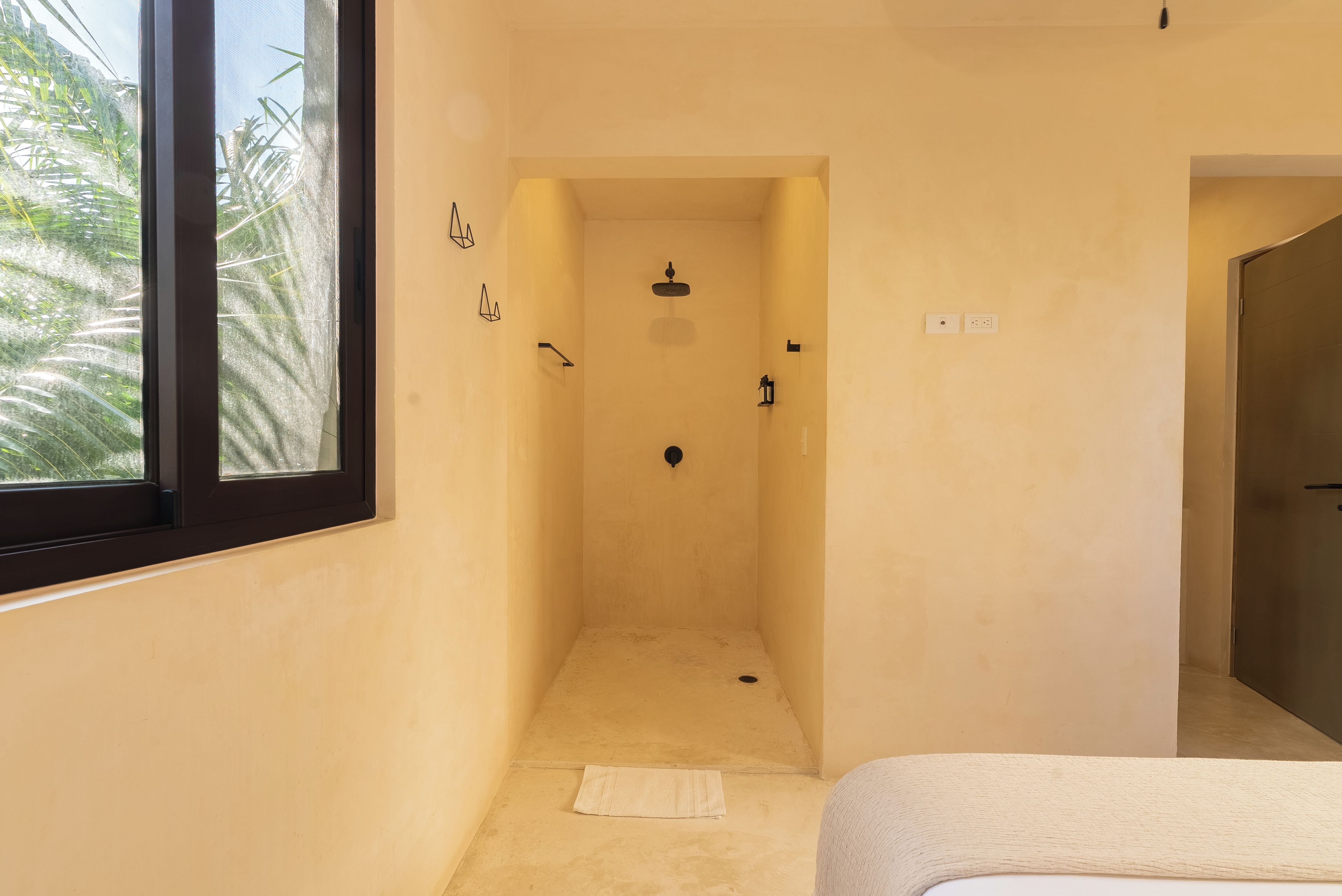 A narrow shower room with a window, a towel on the floor, and a door leading to another room.