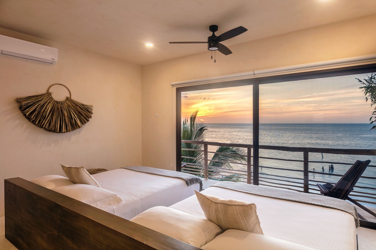 A bedroom with two beds, a ceiling fan, and a large window overlooking the sea.