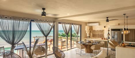 A modern living room with a large window overlooking the ocean, wicker furniture, and a kitchen area.