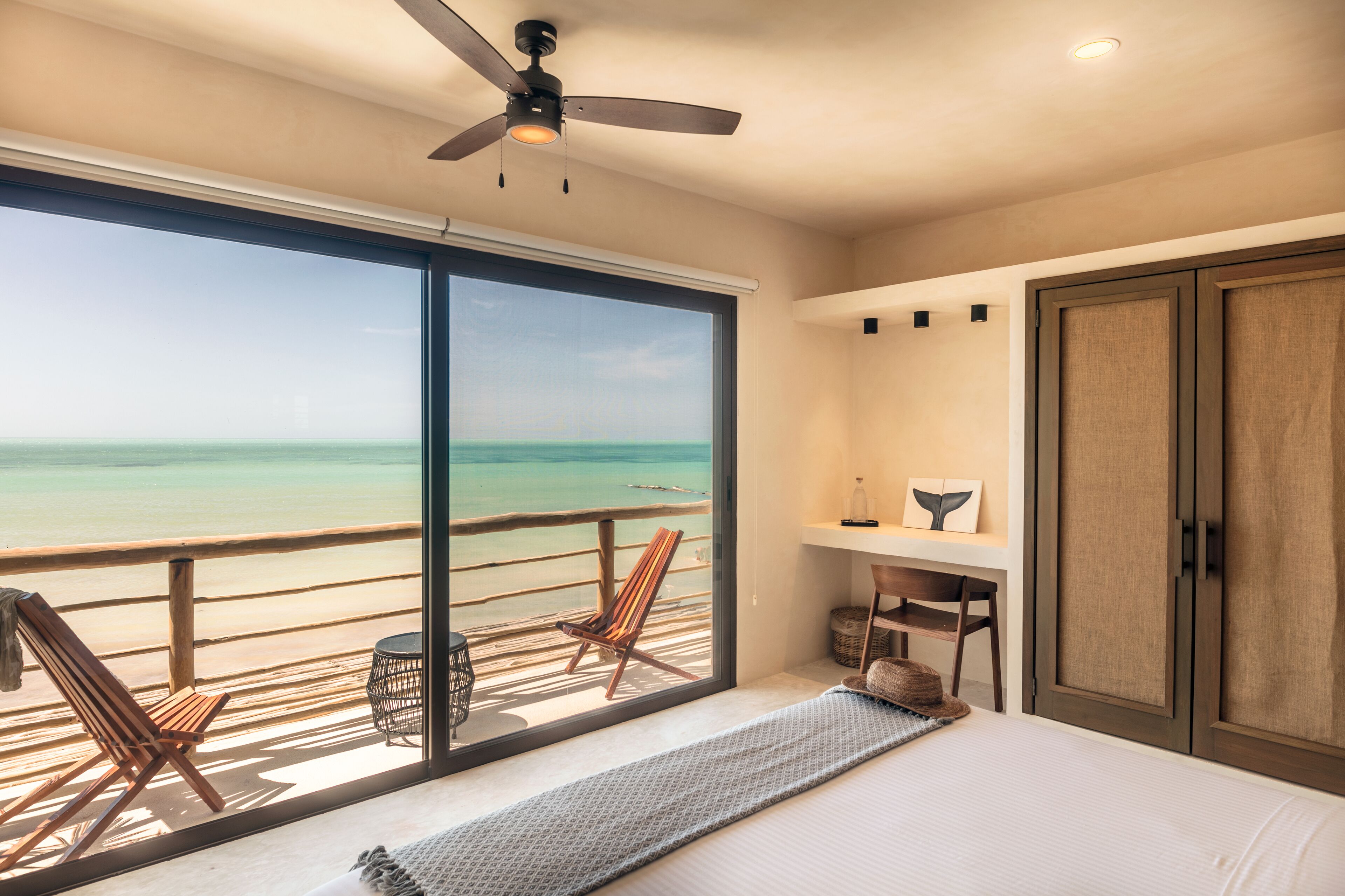 Un dormitorio con un ventanal grande que da a una playa, un ventilador de techo y una cama con vistas.