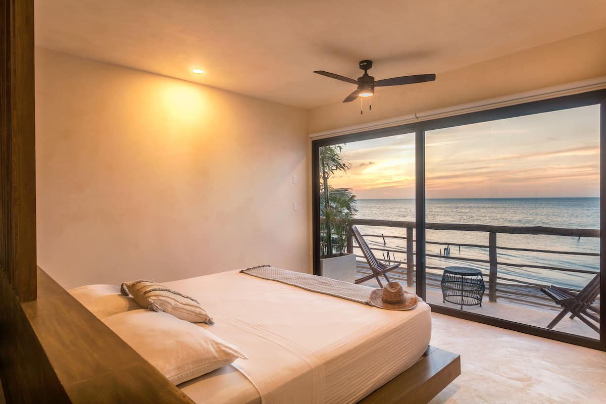 Un dormitorio con un ventanal grande con vistas a la playa al atardecer, un ventilador de techo y una cama con almohadas y un sombrero.
