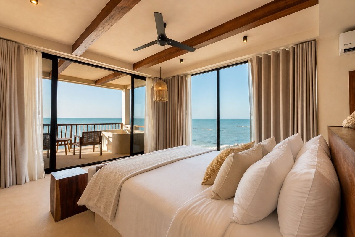 A bedroom with a large bed, a ceiling fan, and a view of the sea through a sliding glass door.