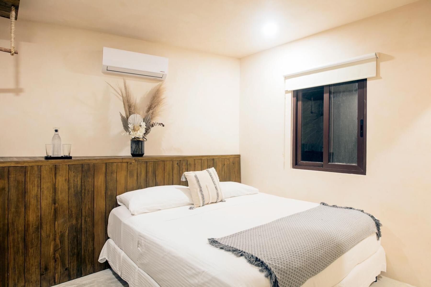 Un dormitorio con cabecera de madera, cama con ropa blanca, ventana con marco oscuro y un equipo de aire acondicionado en la pared.